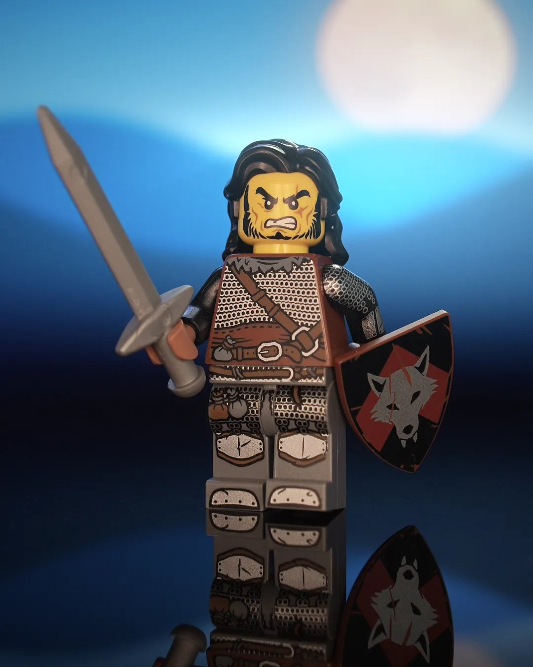 Castle Clad Bricks pad-printed Wolfpack minifig with long black hair, yellow head, fierce grimace, scars and black chinstrap beard. Reddish-brown torso with dark-bluish-gray fur collar, dagged chainmail and medium-brown strap and belt; black arms with chainmail and splinted forearms. Dark-bluish-gray legs with mail skirt, loot bags, iron-clad feet and medium-brown trimmed knee guards. Holds a flat-silver longsword and a reddish-brown heater shield with a silver wolf head on a red upward chevron over black.