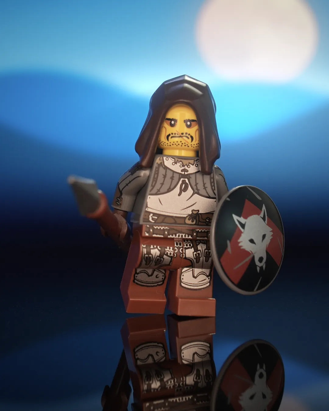 Castle Clad Bricks pad-printed Wolfpack minifig in a dark-brown open hood, with yellow head, stern scowl, stubble, mustache and lip scar. Dark-bluish-gray torso with looted plate: gorget and plackart laced over gambeson, dark-tan fur collar and pouch, and long dark-brown leather gauntlets. Reddish-brown legs with segmented poleyns, short mail skirt and tasset plates on dark-brown straps. Holds a spear and a dark-bluish-gray round shield with a silver wolf head on a dark-red upward chevron over black.