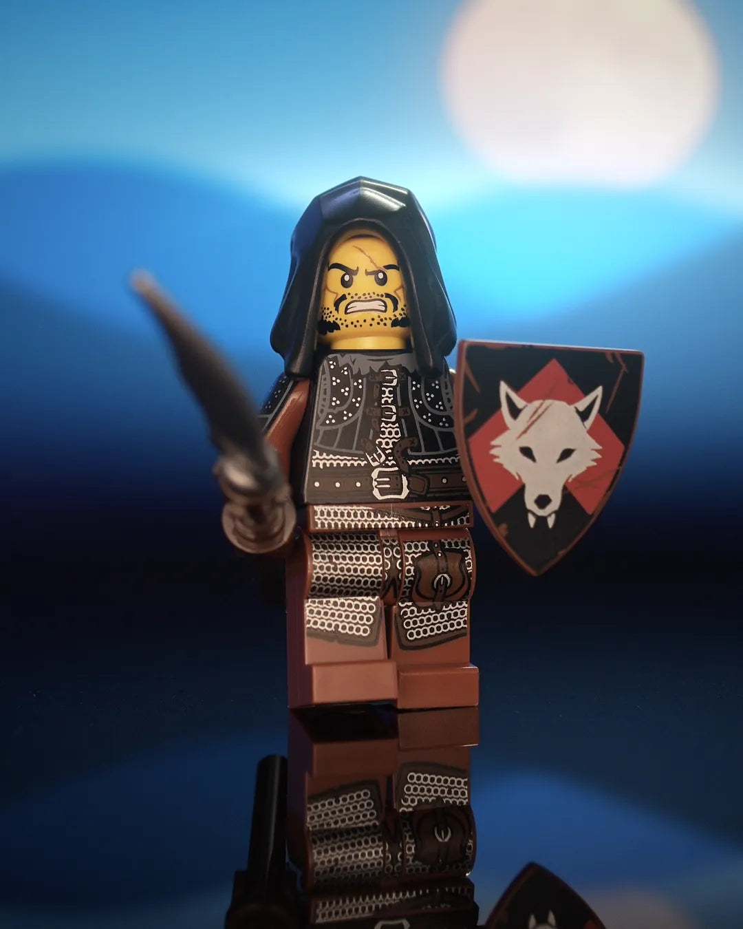 Castle Clad Bricks hooded bandit minifig in a black open hood, with a scarred, snarling, stubbled face. He wears a black studded brigandine vest with fur collar, pauldrons and a heavy buckled belt, reddish-brown arms, and reddish-brown legs printed with silver chainmail, dark-brown leather details and a buckled hip pouch. He holds a glaive with a silver blade and a reddish-brown heater shield with a silver wolf head on a red upward chevron over black.