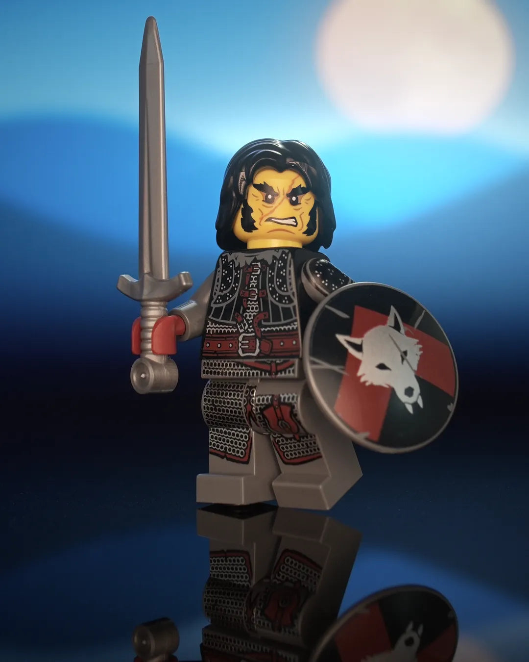 Castle Clad Bricks bandit minifig in a black shoulder length hair, with a scarred face, frown and wild mutton-chops beard. He wears a black studded brigandine vest with fur collar, pauldrons and a heavy buckled belt, dark-bluish-grey arms, and dark-bluish-grey legs printed with silver chainmail, dark-red leather details and a buckled hip pouch. He holds a flat-silver greatsword and a dark-bluish-grey round shield with a silver wolf head on a dark-red upward chevron over black.
