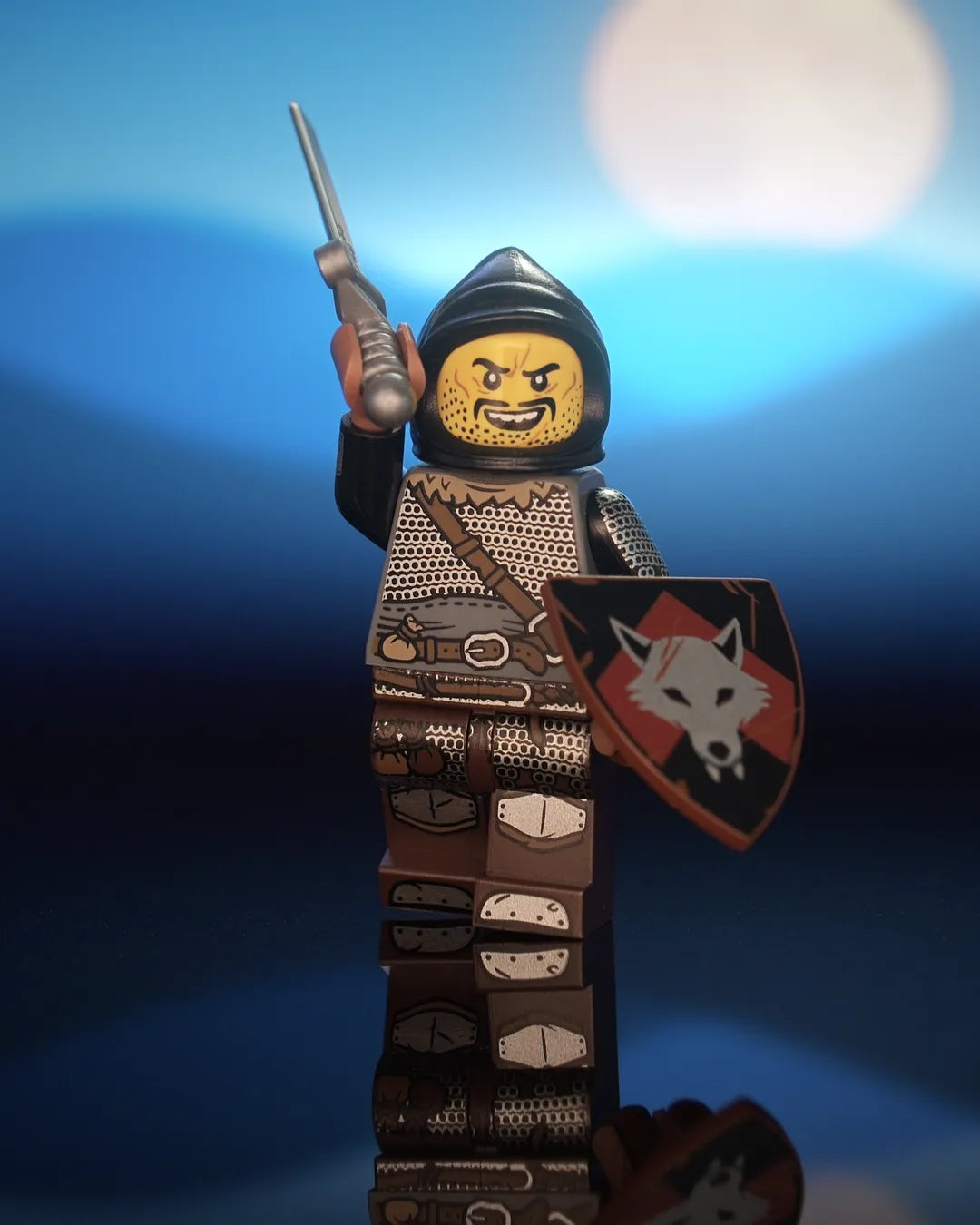 Castle Clad Bricks pad-printed Wolfpack minifig in a black farmer's cowl, with yellow head, evil smile, chin scar, stubbled beard and mustache. Dark-bluish-gray torso with dark-tan fur collar, dagged chainmail and medium-brown strap and belt; black arms with chainmail and splinted forearms. Dark-brown legs with mail skirt, loot bags, iron-clad feet and medium-brown trimmed knee guards. Striking pose with a shortsword; reddish-brown heater shield with a silver wolf head on a red upward chevron over black.