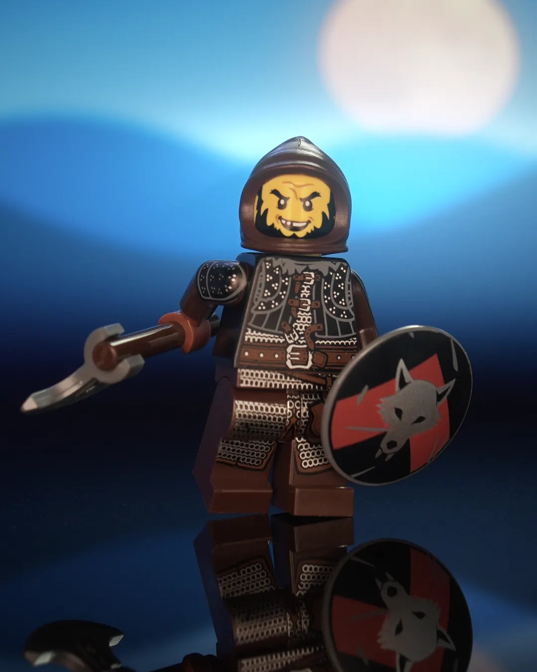 Castle Clad Bricks bandit minifig in a dark-brown farmers cowl, with a wrinkled forehead, crooked smile and wild chinstrap beard. He wears a black studded brigandine vest with fur collar, pauldrons and a heavy buckled belt, dark-brown arms, and dark-brown legs printed with silver chainmail, reddish-brown leather details and a buckled hip pouch. He holds a hand-axe with a silver blade and a dark-bluish-grey round shield with a silver wolf head on a red upward chevron over black.